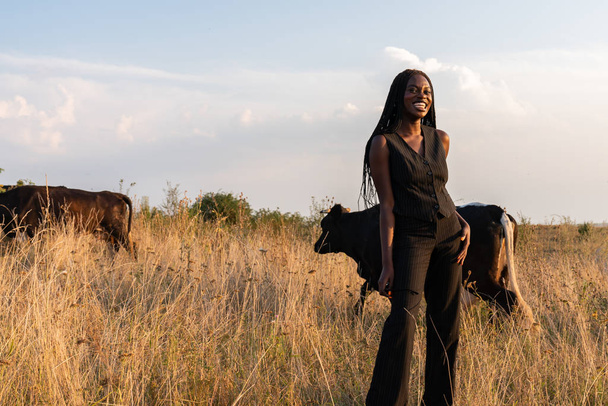 Feliz chica africana sonriente en ropa negra se encuentra entre el campo, las vacas pastan en el fondo
 - Foto, imagen