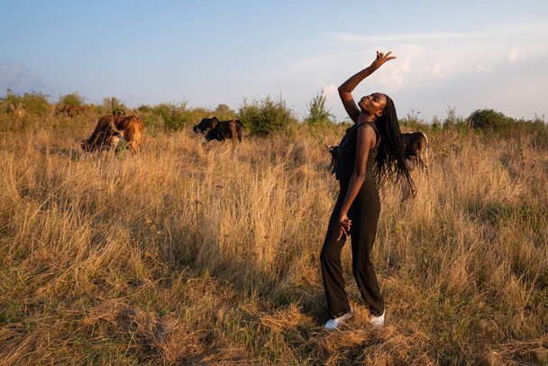Jovem menina africana feliz em roupas pretas dança entre o campo de grama seca, vacas pastam no fundo
 - Foto, Imagem