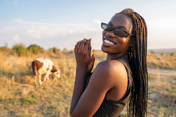 Feliz niña africana sonriente en gafas de sol negras se encuentra entre el campo y mirando a la cámara sobre su hombro, vaca pastando en el fondo
 - Foto, imagen