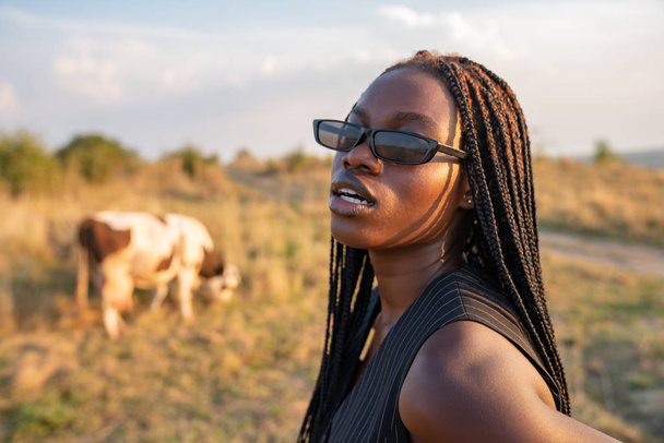Close up retrato da jovem menina africana em colete preto entre o campo, vaca pastam no fundo
 - Foto, Imagem