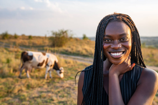 Κοντινό πορτραίτο της νεαρής Αφρικάνας με το μαύρο γιλέκο ανάμεσα στο χωράφι, η αγελάδα βόσκει στο βάθος - Φωτογραφία, εικόνα