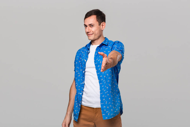 Красивый мужчина в белой футболке и голубой рубашке предлагает руку для рукопожатия изолированы на сером фоне
. - Фото, изображение