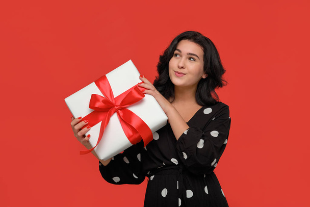 Mujer morena en un vestido negro sonriendo sosteniendo la caja de regalo de Navidad de color blanco con cinta roja
. - Foto, Imagen