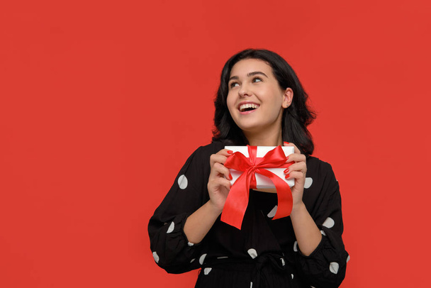 brünette Frau in einem schwarzen Kleid lächelnd mit Weihnachtsgeschenkbox weißer Farbe mit roter Schleife. - Foto, Bild