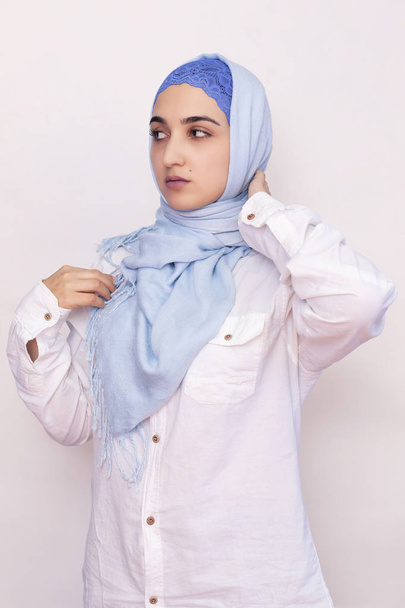 Elegancka muzułmanka w białej koszuli i jasnoniebieskim hidżabie. Stylowa irańska dziewczyna w muzułmańskim ubraniu. Odosobniony portret atrakcyjnej kobiety z Bliskiego Wschodu - Zdjęcie, obraz