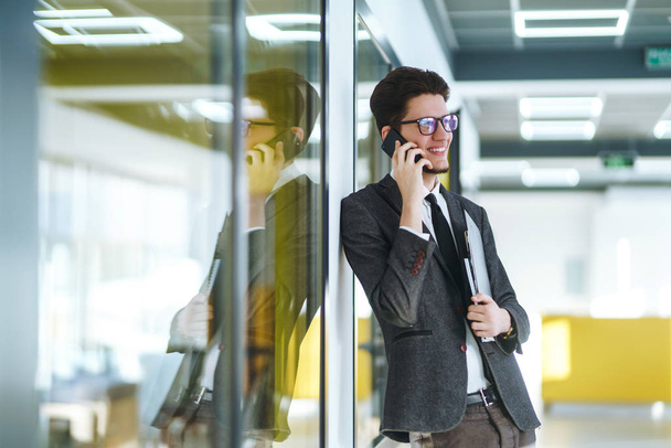 Young businessman in glasses talking on cell phone standing in the modern office. Stylish man in a suit with folder standing and talking on telephone. Communication concept. - Foto, Bild