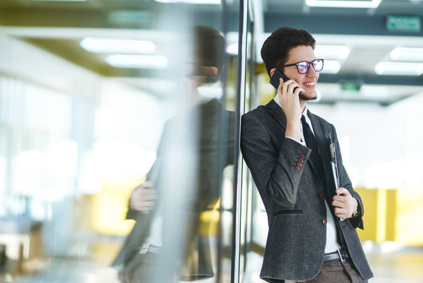 Young businessman in glasses talking on cell phone standing in the modern office. Stylish man in a suit with folder standing and talking on telephone. Communication concept. - Foto, imagen
