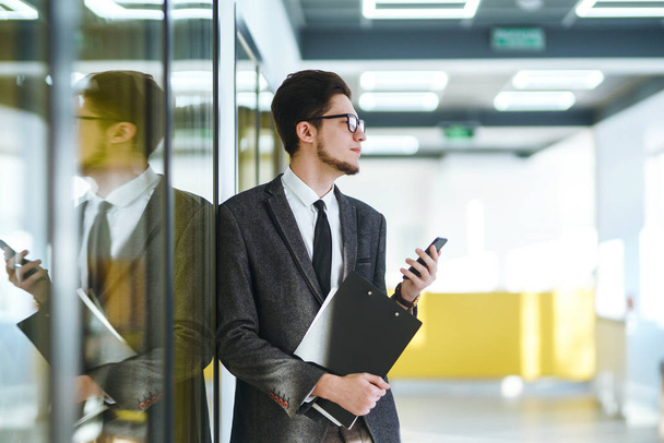 Young office worker in glasses using mobile smart phone. Businessman holds telephone in hand. Stylish man in a suit with phone in hands prints a message on the phone.  Communication concept. - Foto, imagen