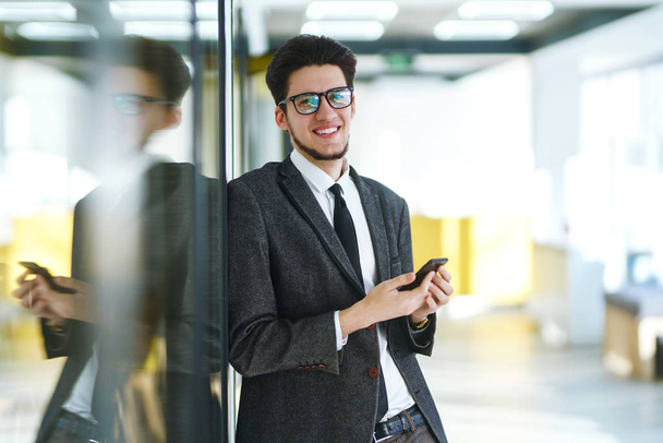 Businessman with phone in hands smiling and looking at the camera. Stylish man in a suit with phone in hands  standing in the modern office. Young office worker in glasses using mobile smart phone. - Φωτογραφία, εικόνα