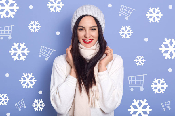 Μελαχρινή γυναίκα με όμορφα μαλλιά φοράει ζεστό χειμερινό καπέλο, sacarf και λευκό πουλόβερ, φαίνεται χαρούμενος, κρατά τα χέρια στο λαιμό της, θηλυκό με φωτεινά χείλη στέκεται ενάντια σε μπλε τοίχο στούντιο με νιφάδες χιονιού. - Φωτογραφία, εικόνα