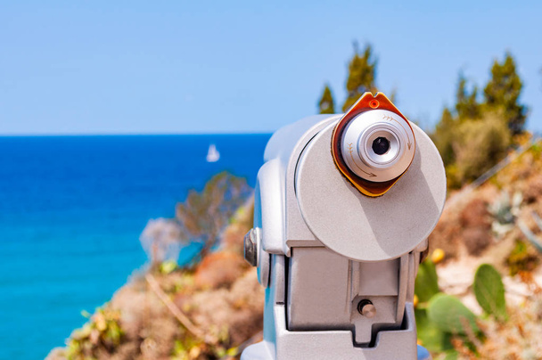 Stacjonarny teleskop turystyczny wykonany z metalu na jednej nodze stojący na krawędzi klifu w pobliżu drewnianego płotu kłód i obserwujący tyrreńskie morze z pływającym białym jachtem żaglowym w Tropei - Zdjęcie, obraz