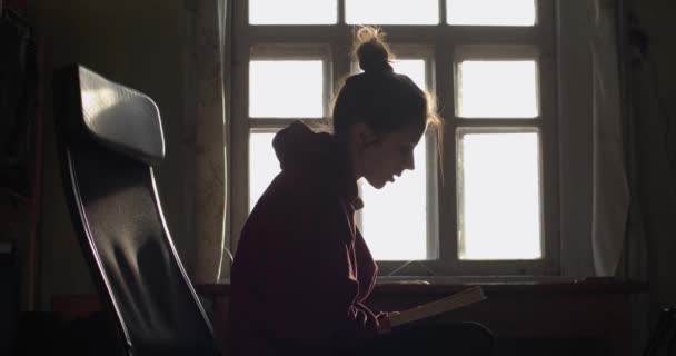 Side muotokuva tyttö pulla lukeminen paperikirja ääneen sisätiloissa vintage maan ikkunanpuitteet taustavalaistu. Nuori nainen istuu nojatuolissa auringonvalossa luonnonvalossa hidastettuna. Hidas elämäntapa
 - Materiaali, video