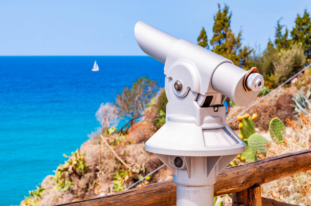 木製の丸太垣の近くの崖の端に立っている1本の脚に金属製の固定観光望遠鏡とトロピープに浮かぶ白いセーリングヨットでティレニア海を観察します - 写真・画像
