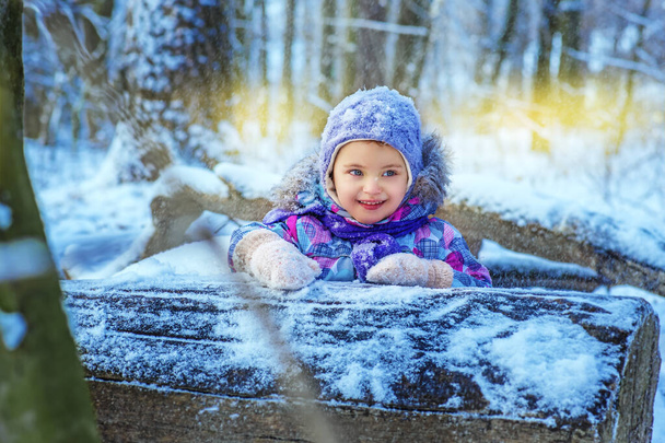 Mała 4-letnia staruszka w śnieżnym lesie zimowym gra na świeżym powietrzu. Dziecko w kolorowych ubraniach uśmiecha się i odwraca wzrok. Drewniany kufer na oczach dziewczyny. Zachód słońca - Zdjęcie, obraz
