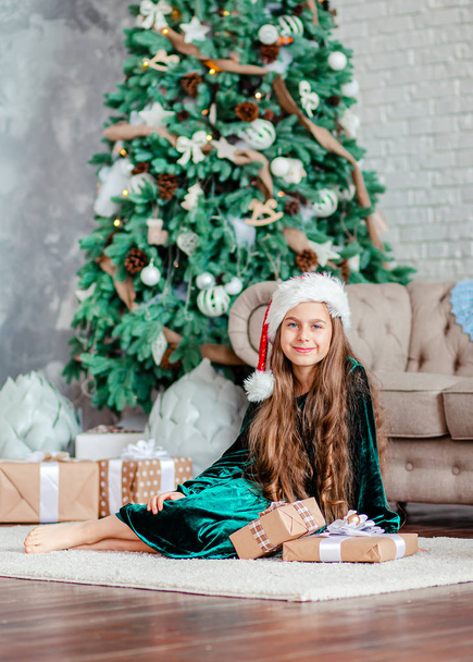 Little girl Santa Claus hat with gifts under Christmas tree sitting by the fireplace, unpacks gifts. - Zdjęcie, obraz