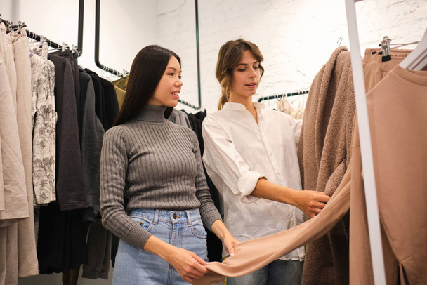 Young designer showing clothes from new collection to attractive Asian girl in modern clothes store - 写真・画像