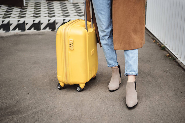 Close up stylish girl in coat standing with yellow suitcase on street - 写真・画像
