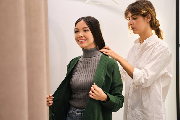Young fashion consultant helping trying on cardigan to pretty positive Asian girl in modern store - 写真・画像