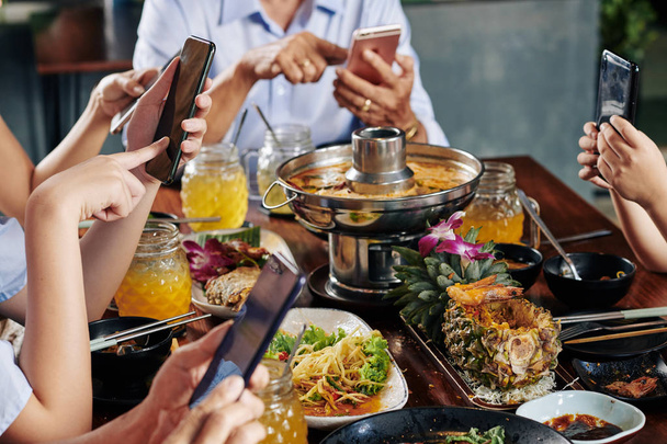 Dłonie gości partii siedzi przy stole z tradycyjną kuchnią azjatycką i SMS-y znajomych sprawdzania mediów społecznościowych na smartfonach - Zdjęcie, obraz