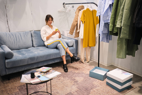 Νέοι κομψό σχεδιαστής μόδας με αυτοπεποίθηση χρησιμοποιώντας το κινητό τηλέφωνο στον καναπέ στο σύγχρονο εκθεσιακό χώρο - Φωτογραφία, εικόνα