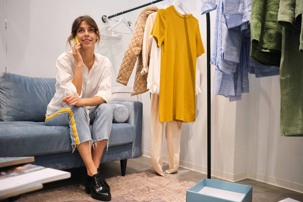 Mladý ležérní módní návrhář šťastně mluví na mobilu na pohovce v moderní showroom - Fotografie, Obrázek