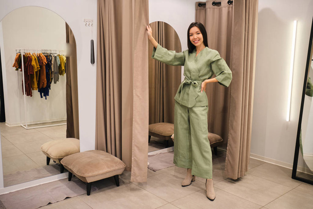 Atractiva chica asiática alegre probándose un traje elegante mirando felizmente en la cámara en el vestidor de la tienda de ropa moderna
 - Foto, Imagen