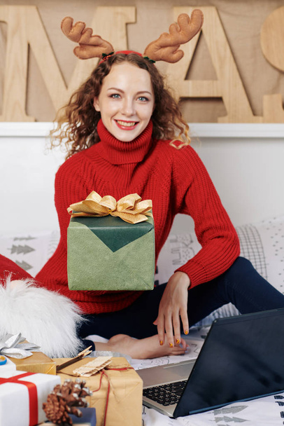 Щаслива молода жінка простягає руку з різдвяним подарунком, загорнутим в зелений папір
 - Фото, зображення