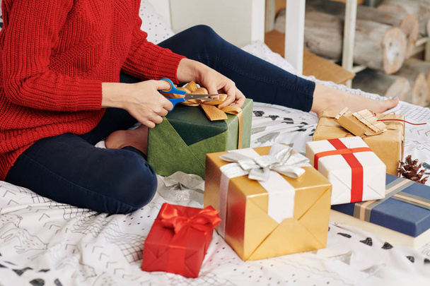 Cropped image of young woman sitting on bed and cutting ribbon on wrapped Christmas present - 写真・画像