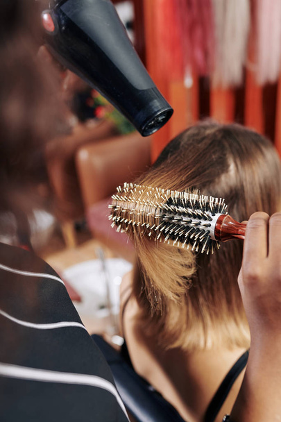 Професійний перукар з використанням круглої щітки та сушарки при сушінні волосся жіночого клієнта в салоні краси
 - Фото, зображення
