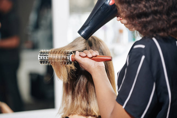 Ammattimainen stylisti harjaus ja hiustenkuivaaja hiukset naispuolinen asiakas, näkymä takaa
 - Valokuva, kuva