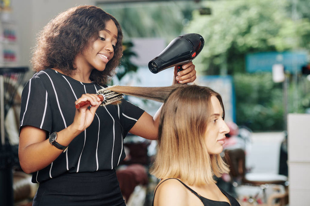 Pozitif genç siyah kuaför güzellik salonunda çalışmaktan ve müşterinin saçlarını kurutmaktan zevk alıyor. - Fotoğraf, Görsel