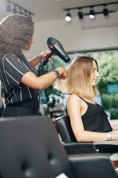 Стилист, использующий профессиональную сушилку при сушке волос молодой красивой женщины в ее салоне
 - Фото, изображение