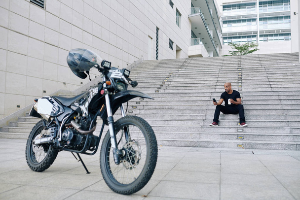 Yakışıklı kaslı motosikletçi basamaklarda oturuyor, içiyor kahveyi götürüyor ve sosyal medyayı kontrol ediyor. - Fotoğraf, Görsel