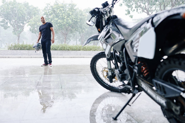 Feliz homem sorridente com capacete na mão olhando para sua garagem aberta com motocicleta
 - Foto, Imagem