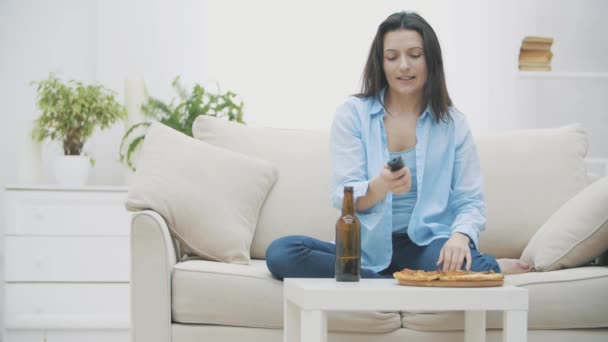 A young woman is sitting on a sofa, eating pizza and drinking beer. She is turning TV on. Yummy-yummy. 4K. - Imágenes, Vídeo