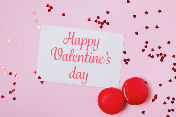 Composición del día de San Valentín con tarjeta de felicitación con texto, dos macarrones y confeti en forma de corazón sobre fondo rosa
 - Foto, imagen