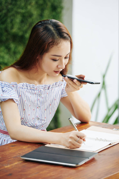 Prety young Asian business lady recording voice message for colleorker and taking notes when sitting at cafe table
 - Фото, изображение