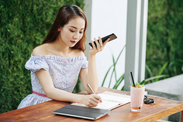 Jeune femme vietnamienne écoutant un message vocal sur son smartphone et prenant des notes dans le planificateur
 - Photo, image