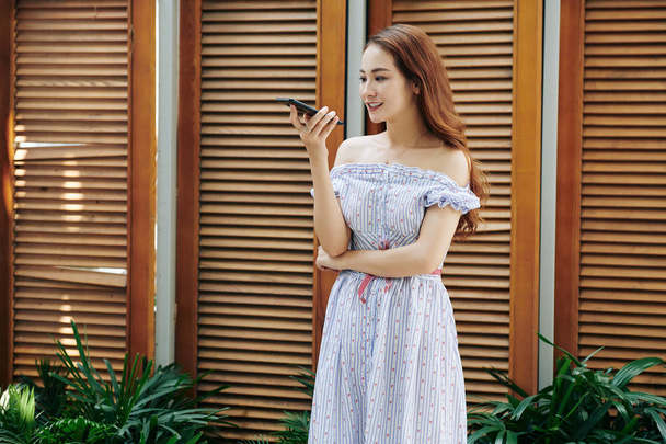 Piękna młoda Azjatka z długimi włosami stojąca na zewnątrz w letniej sukience i nagrywająca wiadomość głosową dla przyjaciela - Zdjęcie, obraz