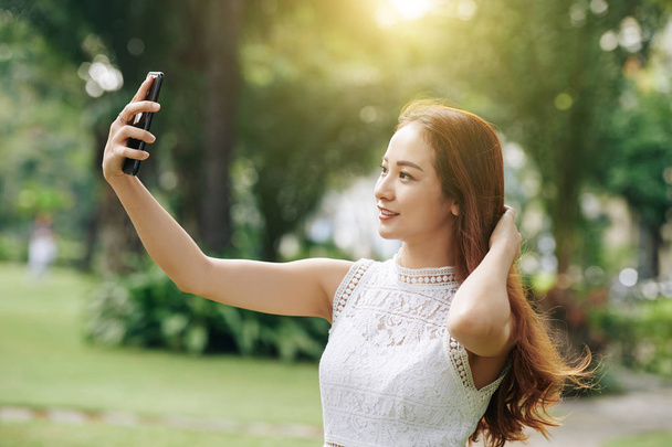 Όμορφη νεαρή βιετναμέζα γυναίκα λαμβάνοντας selfie στο smartphone όταν περπατάτε στο πάρκο - Φωτογραφία, εικόνα
