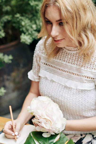 Young beautiful blond woman with hydrangea flower writing in her diary - Photo, Image