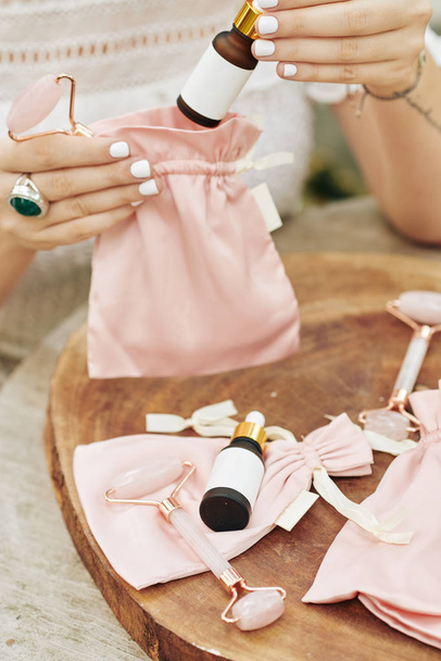 Close-up beeld van kleine cosmetica bedrijf eigenaar verpakking set van verjongende essentie en kwarts massage roller in zijde roze zakken - Foto, afbeelding