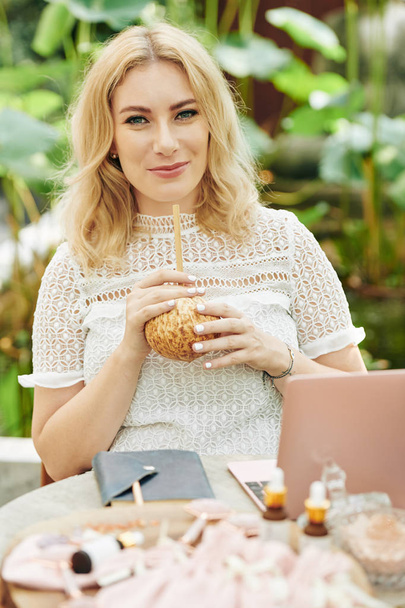 glückliche charmante junge Frau mit Kokosmilch, wenn sie am Cafétisch am Businessplan für ihr Kosmetikgeschäft arbeitet - Foto, Bild