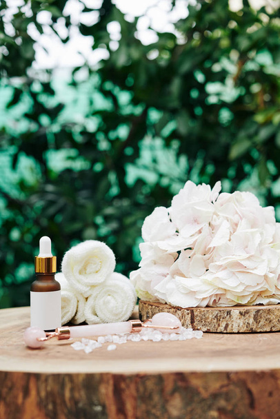 Bottle of moisturizing facial serum and quartz massage roll on wooden stump with blooming flower and small rolled towels - Photo, Image