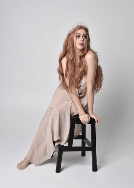 full length portrait of a  girl wearing a nude flowy dress and crystal crown. Sitting on a chair against a grey studio background - Φωτογραφία, εικόνα