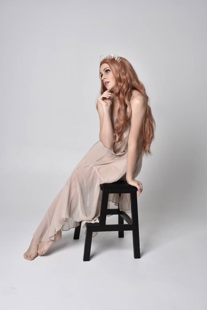 full length portrait of a  girl wearing a nude flowy dress and crystal crown. Sitting on a chair against a grey studio background - Foto, imagen
