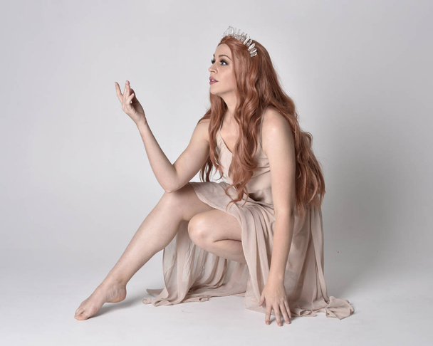 full length portrait of a pretty, fairy girl wearing a nude flowy dress and crystal crown. Seated pose against a grey studio background. - Valokuva, kuva