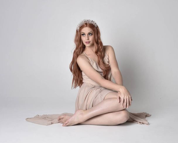 full length portrait of a pretty, fairy girl wearing a nude flowy dress and crystal crown. Seated pose against a grey studio background. - 写真・画像