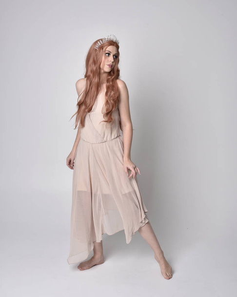 Ganzkörperporträt eines hübschen, feenhaften Mädchens in einem nackt flowy Kleid und Kristallkrone. Stehen und Tanzen vor grauem Studiohintergrund. - Foto, Bild