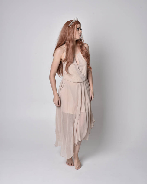 Ganzkörperporträt eines hübschen, feenhaften Mädchens in einem nackt flowy Kleid und Kristallkrone. Stehen und Tanzen vor grauem Studiohintergrund. - Foto, Bild